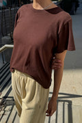 A person wearing a short-sleeve crewneck t-shirt in a muted brownish-red color, paired with light beige drawstring pants, standing outdoors on a sunny day.