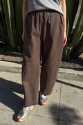 A person wearing loose brown wide-leg pants with a gray top and metallic silver sandals, standing outdoors on a concrete sidewalk in front of a cactus plant.