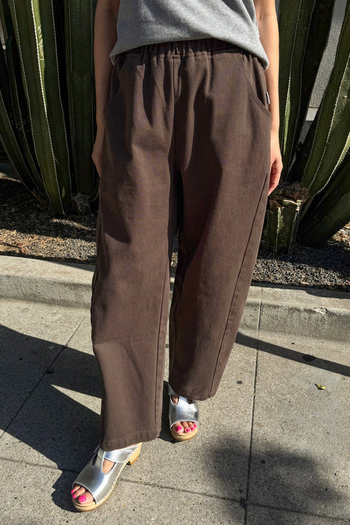 A person wearing loose brown wide-leg pants with a gray top and metallic silver sandals, standing outdoors on a concrete sidewalk in front of a cactus plant.