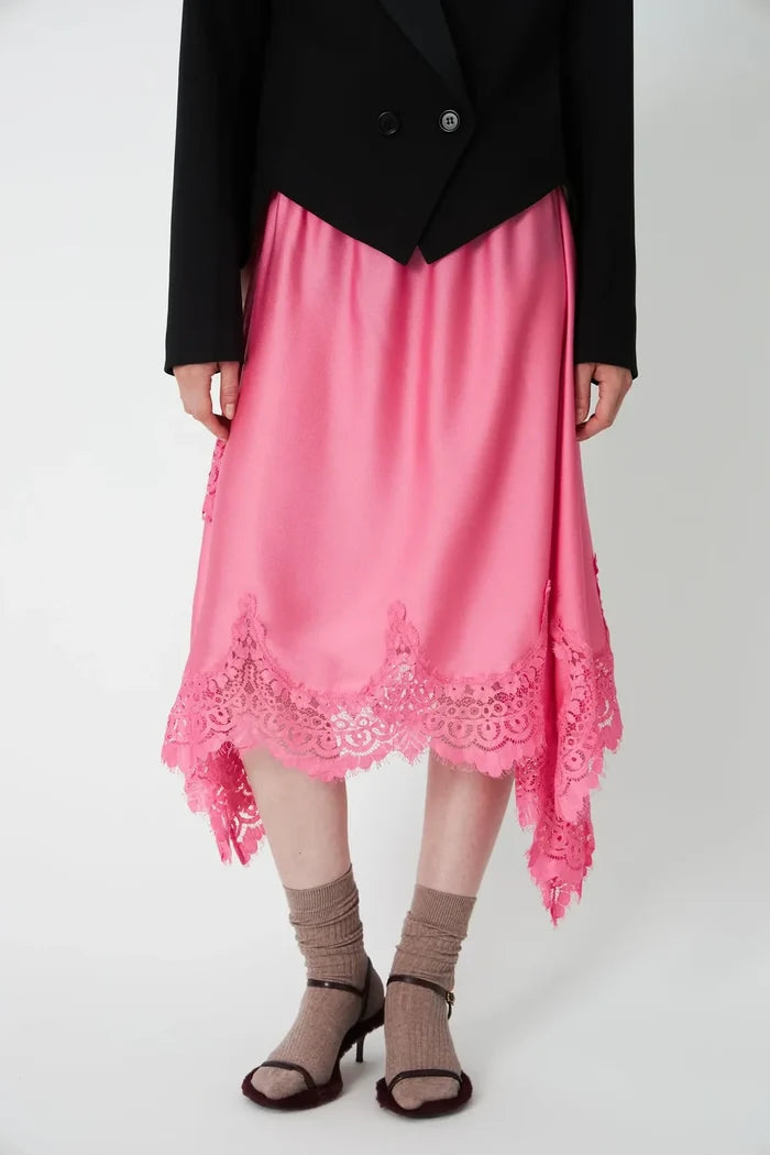 A pink high-low midi skirt with a scalloped lace hem, styled with a black blazer, beige socks, and brown heeled sandals against a light gray studio background.