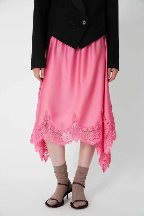 A pink high-low midi skirt with a scalloped lace hem, styled with a black blazer, beige socks, and brown heeled sandals against a light gray studio background.