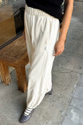 A person wearing loose, cream-colored pants with a small side pocket, paired with a black top and dark shoes, standing on a concrete floor next to a rustic wooden stool.