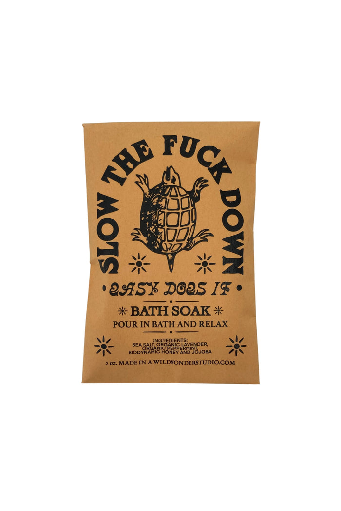 A rectangular kraft paper bath soak packet with black printed text and a turtle illustration, reading 'SLOW THE FUCK DOWN'.