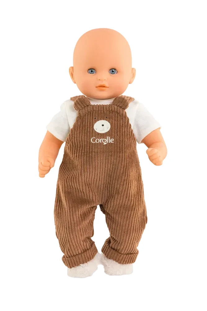 A baby doll with a bald head and blue eyes, wearing brown corduroy overalls over a white t-shirt. The overalls feature a white embroidered bear face and the name 'corolle' in white script.