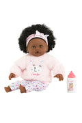 A Corolle brand doll with dark skin and curly black hair, wearing a pink fuzzy long-sleeve top and matching patterned pants, sitting next to a pink baby bottle and pacifiers on a white background.