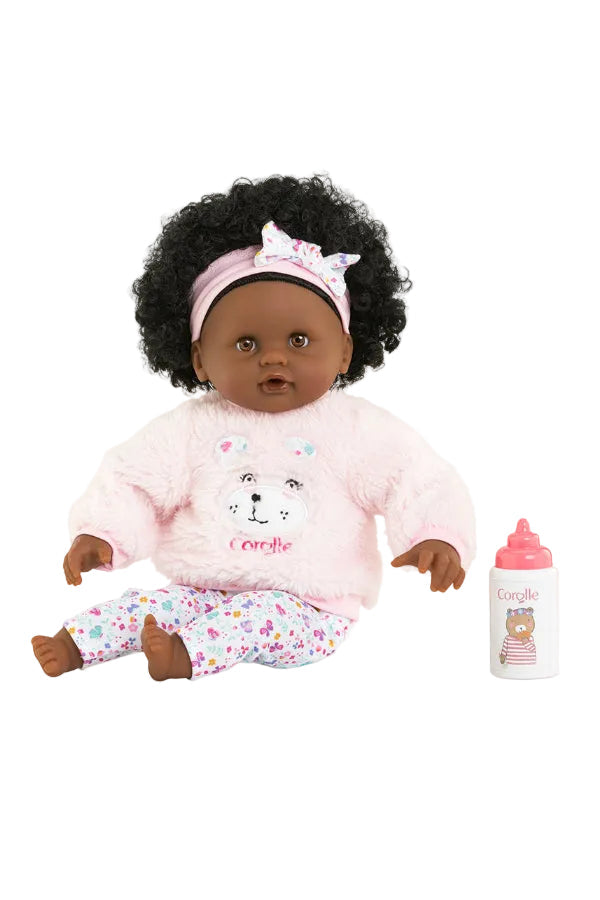 A Corolle brand doll with dark skin and curly black hair, wearing a pink fuzzy long-sleeve top and matching patterned pants, sitting next to a pink baby bottle and pacifiers on a white background.