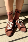 A person wearing brown ribbed crew socks and matching suede low-top sneakers with white laces, standing on a concrete floor in direct sunlight.