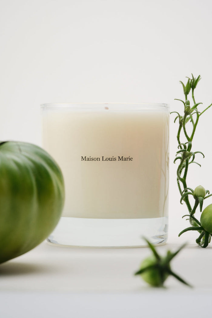 Candle with 'Maison Louis Marie' branding next to a green tomato and plant on a white surface