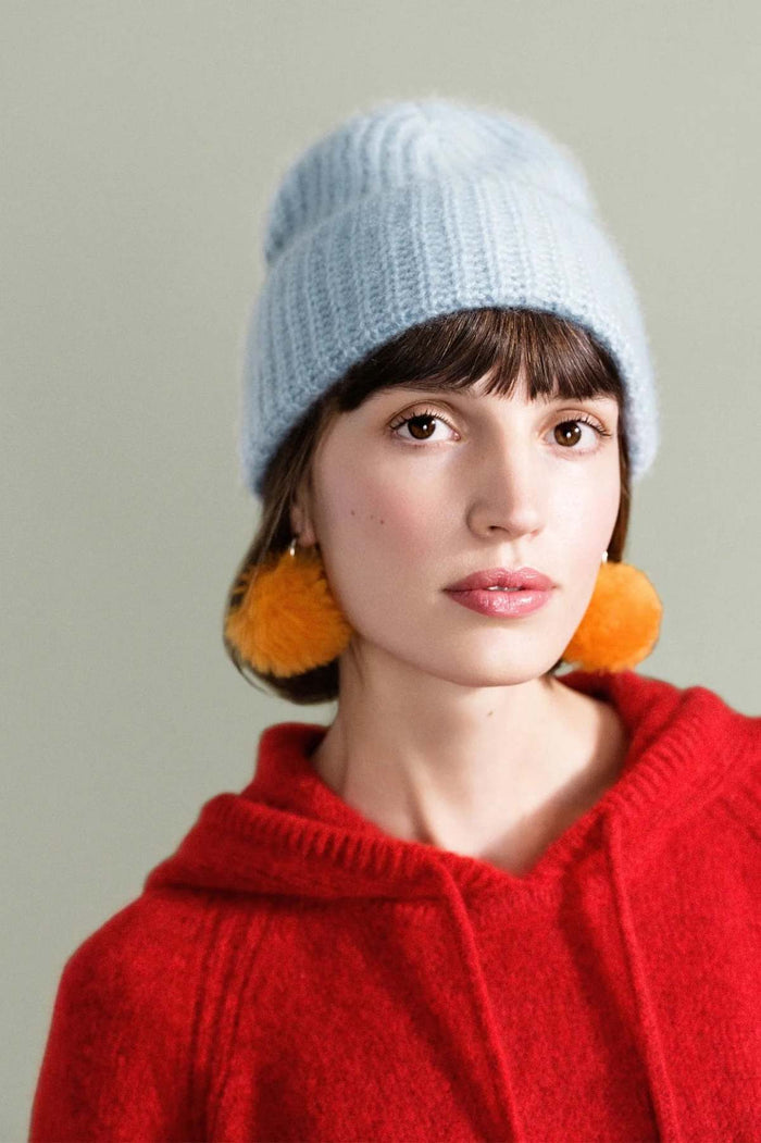 A woman wearing a light blue ribbed knit beanie, large orange pom-pom earrings, and a red knit hoodie against a plain light gray background.