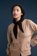 A woman wearing a beige knit cardigan with front buttons and patch pockets, styled with a black neck scarf, set against a dark blue studio background.