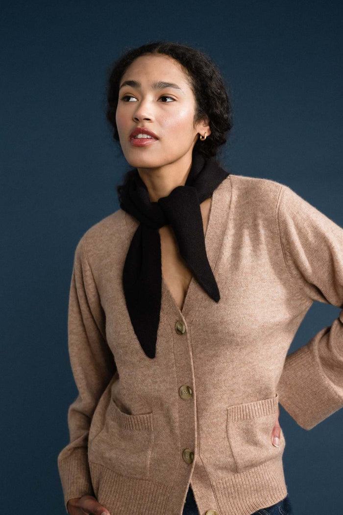 A woman wearing a beige knit cardigan with front buttons and patch pockets, styled with a black neck scarf, set against a dark blue studio background.