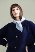 A woman wearing a dark navy blue ribbed knit cardigan sweater with a light blue knotted scarf, posed against a solid light sage green background.