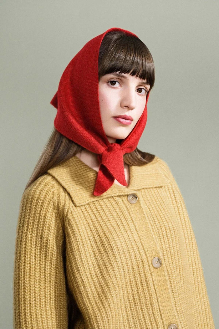 A woman wearing a bright red knitted headscarf and a mustard yellow ribbed cardigan sweater, photographed against a solid light gray background.