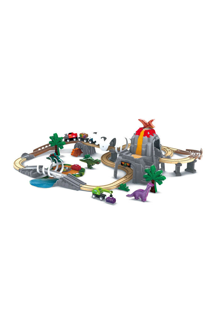 A toy train track set with a gray volcano and dinosaur-themed station, curved tracks, small toy vehicles, palm trees, and a purple dinosaur figure on a plain white background.