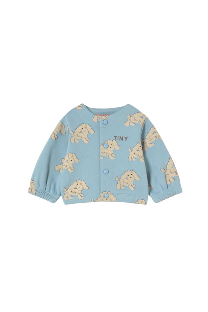 A light blue baby cardigan featuring a repeating beige dog print and the word 'TINY' embroidered on the chest, displayed flat against a warm off-white background.