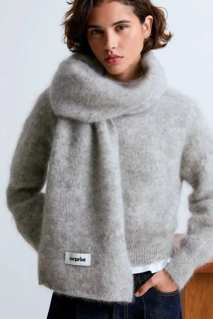 A woman wearing a light gray fuzzy knit sweater with an integrated oversized cowl neck and a matching knit scarf. A small white label reading 'surprise' is visible near the lower left front of the sweater.
