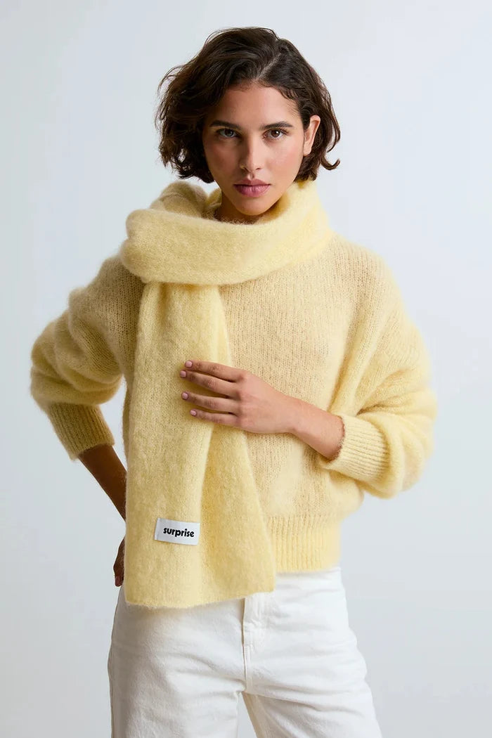 A woman wearing a fuzzy pale yellow knit sweater and matching scarf with a visible 'surplus' label, paired with white pants, against a plain light gray studio background.