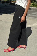 A person outdoors wearing loose black wide-leg pants with a small white side tag, paired with a white top and bright pink heeled thong sandals on a paved street.