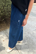 A person wearing wide-leg blue denim pants with a black top and white flats, standing outdoors on a concrete surface next to greenery.