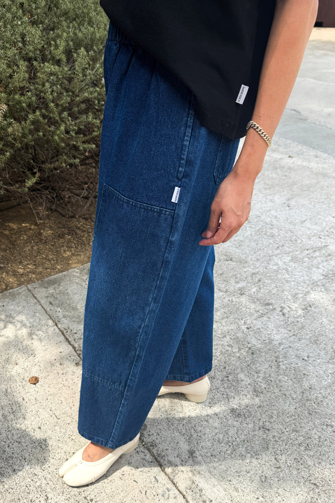 A person wearing wide-leg blue denim pants with a black top and white flats, standing outdoors on a concrete surface next to greenery.