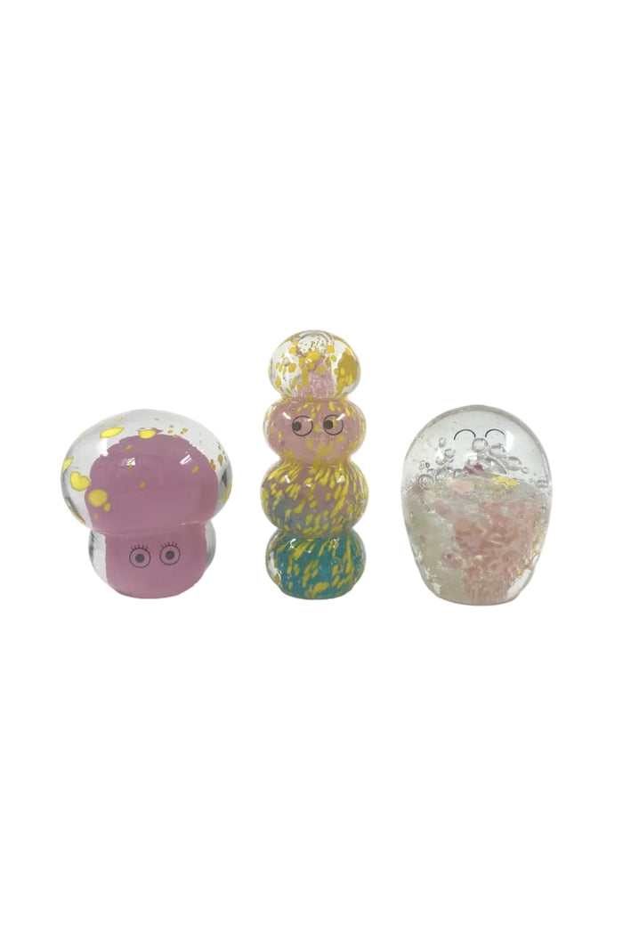 Three small decorative glass figurines on a light gray studio background: a pink mushroom-shaped figure with yellow speckles, a stacked pastel tower with multicolor swirls, and a clear egg-shaped figure filled with pink glitter.