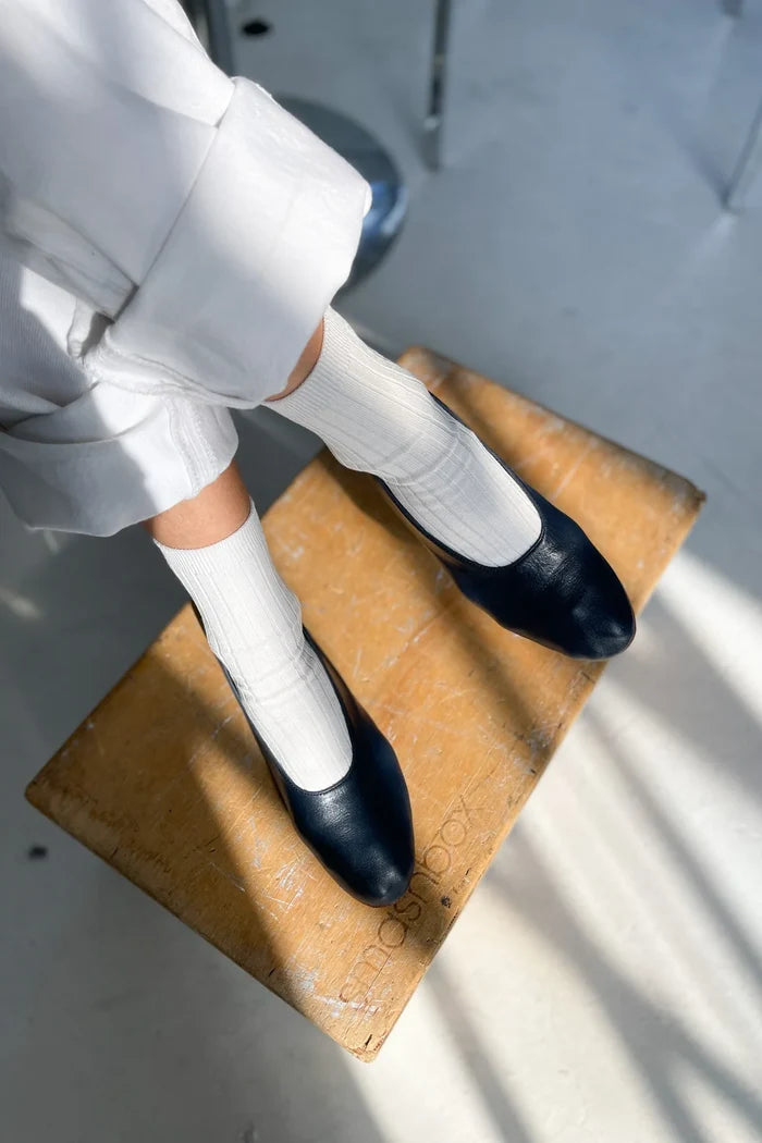 Woman wearing white pants, white socks, and black shoes with her feet against a wooden stool