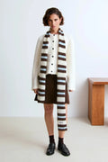 A model stands in a minimalist studio setting wearing a cream-colored cardigan, a brown mini skirt, and a prominent striped fuzzy scarf.