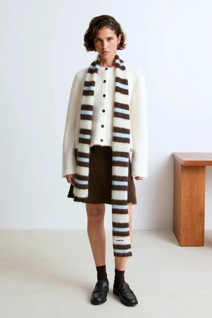 A model stands in a minimalist studio setting wearing a cream-colored cardigan, a brown mini skirt, and a prominent striped fuzzy scarf.
