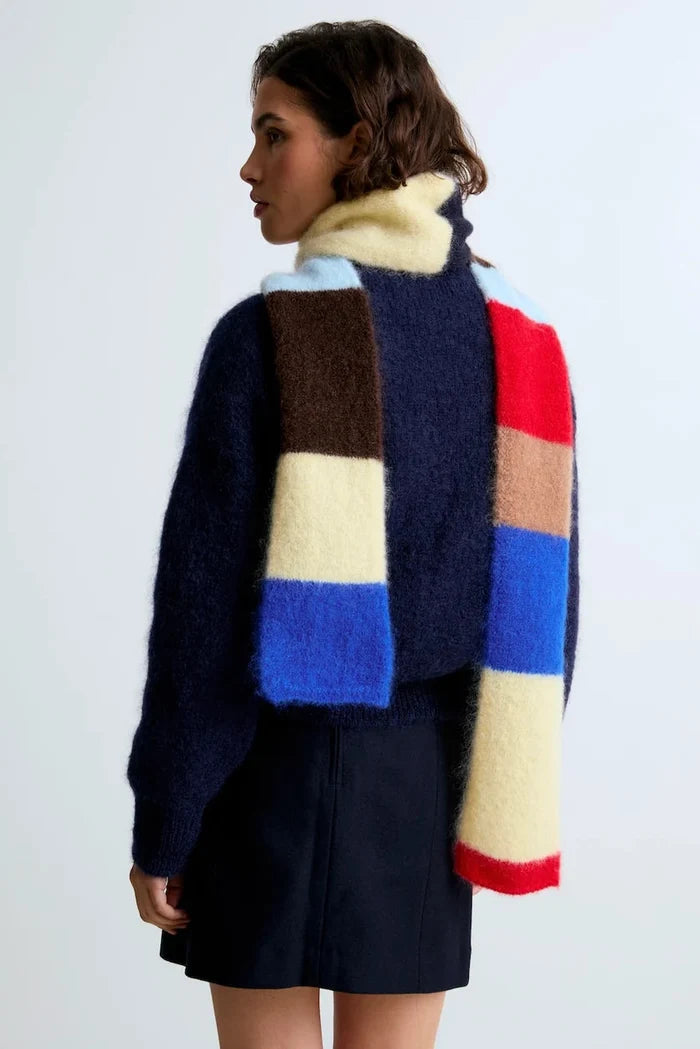 A model shown from the back wearing a fuzzy, multi-colored scarf layered over a navy blue sweater and dark skirt against a light gray studio background.