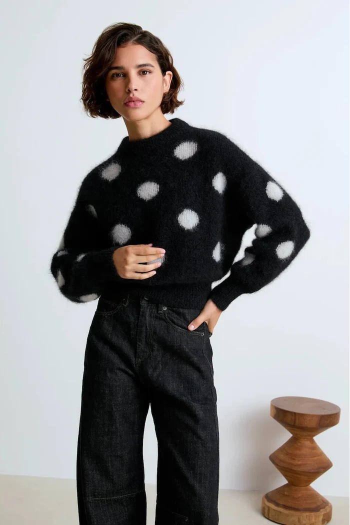 Person wearing a black sweater with white polka dots and dark jeans against a white background.