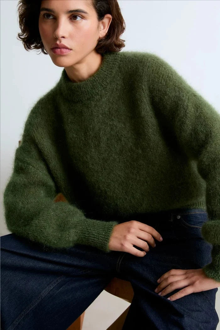 A woman wearing a fuzzy olive green mock-neck sweater and dark blue jeans, seated against a plain light background.