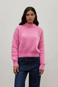 A woman modeling a fuzzy pink mock-neck knit sweater paired with dark blue jeans, standing against a plain light gray studio background.