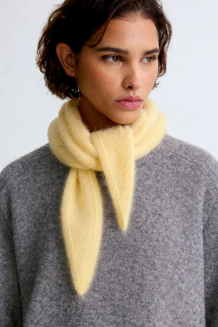 A close-up of a model wearing a soft, pale yellow fuzzy scarf tied around her neck over a heather gray knit sweater, against a plain light gray background.