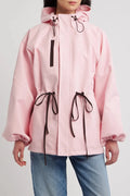 A model wearing a light pink hooded rain jacket with dark brown drawstrings and a vertical zipper pocket, paired with blue jeans against a light gray studio background.