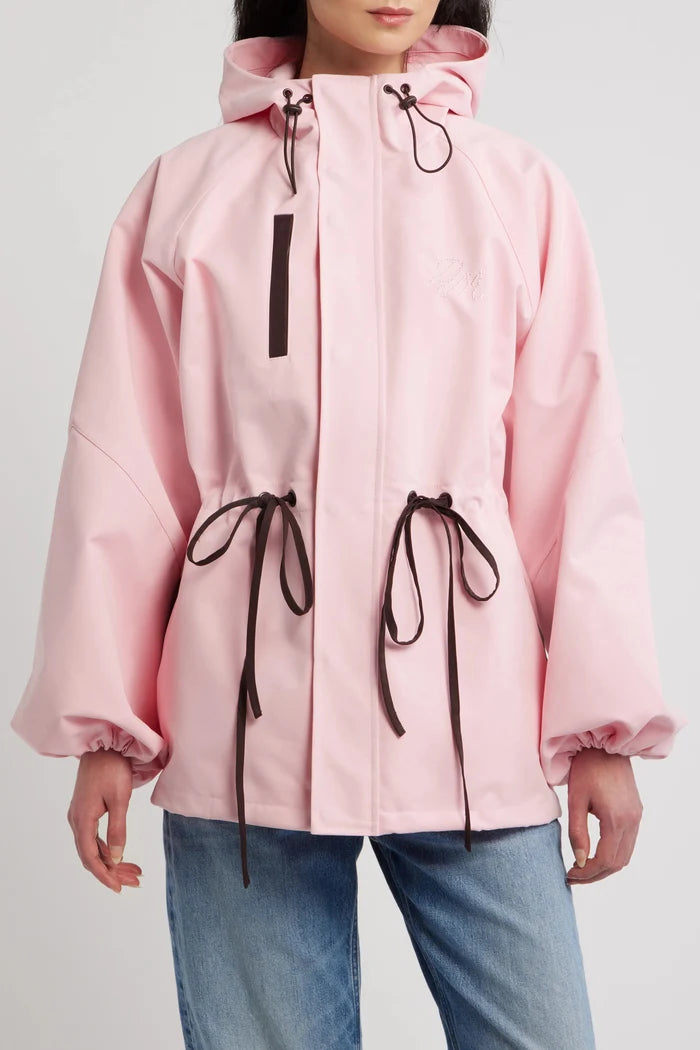 A model wearing a light pink hooded rain jacket with dark brown drawstrings and a vertical zipper pocket, paired with blue jeans against a light gray studio background.