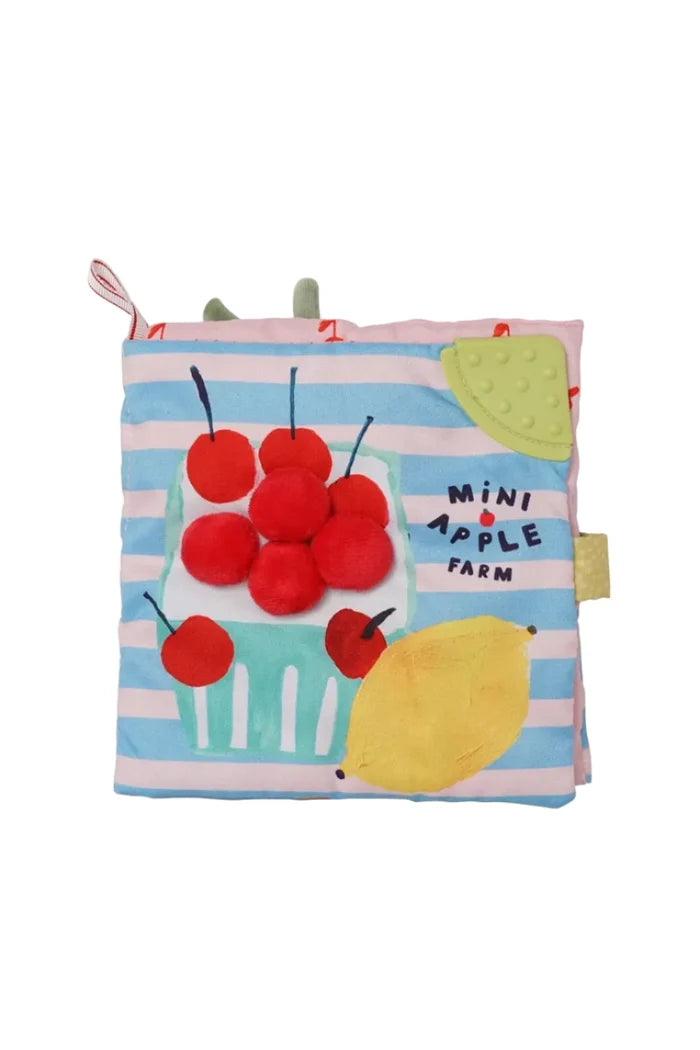 A soft fabric baby book or toy with a striped pink and blue cover, featuring applique illustrations of red cherries, a yellow lemon, a green watermelon slice, and the words 'MINI APPLE FARM'.
