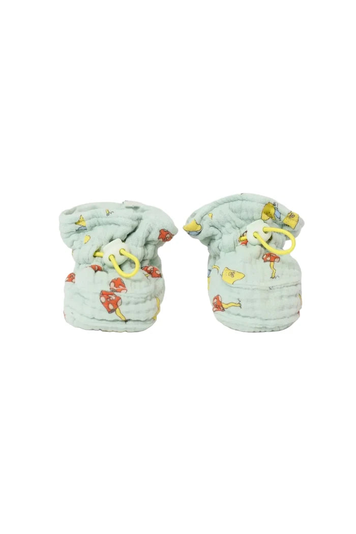 A pair of light sage green muslin baby booties with a colorful cartoon mushroom print and yellow drawstring loops, shown on a plain white background.