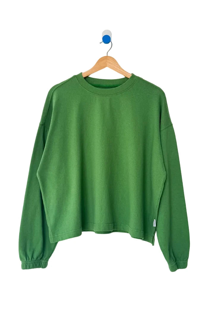 A green long-sleeve cropped sweatshirt hanging on a wooden hanger against a plain white wall.