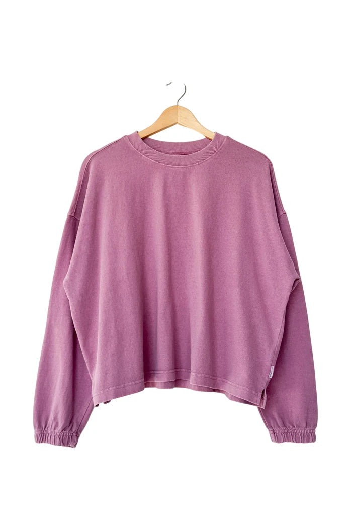 A dusty purple long-sleeve cropped sweatshirt tee on a wooden hanger against a plain white wall.