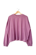 A dusty purple long-sleeve cropped sweatshirt tee on a wooden hanger against a plain white wall.