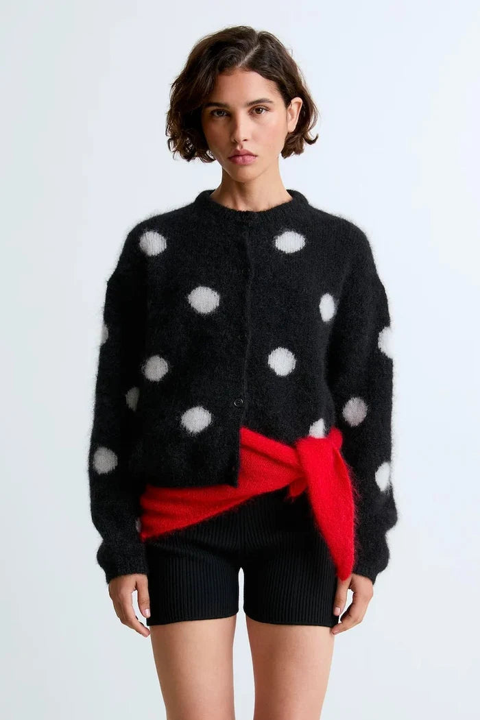 A model wearing a fuzzy black cardigan with white polka dots, styled with a bright red wrap tied around the waist and black ribbed shorts, against a light gray studio background.