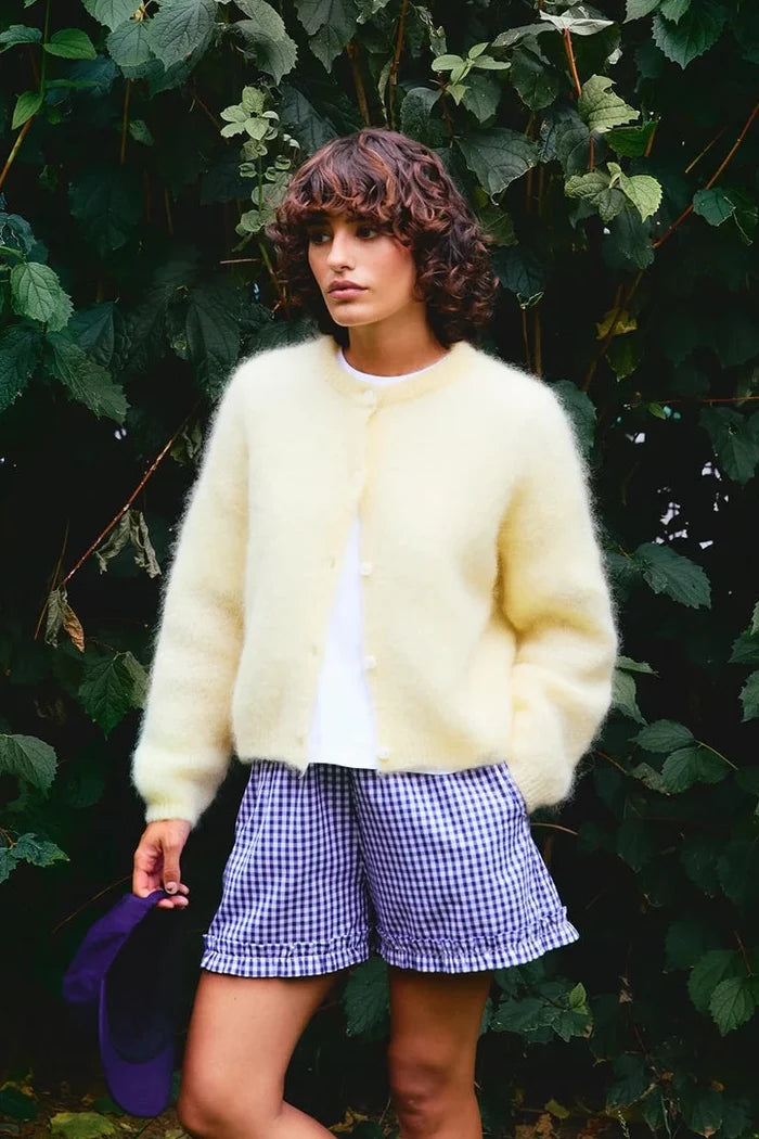 Woman wearing a light yellow cardigan and checkered shorts standing in front of green foliage.