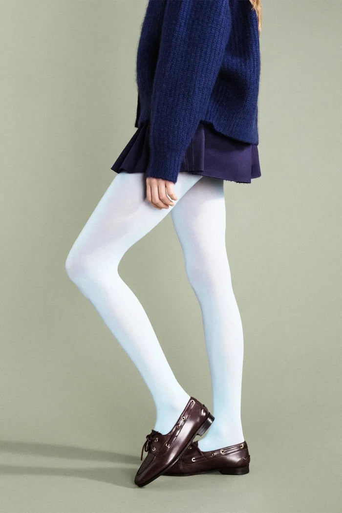 A model wearing white tights, a navy blue sweater, a dark purple pleated skirt, and dark brown leather loafers, shown from the waist down against a solid light gray-green studio background.
