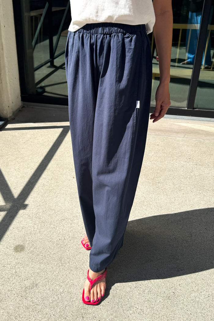 A person wearing loose navy blue drawstring pants and bright pink flip-flop sandals, standing outdoors on a concrete surface near a glass door.