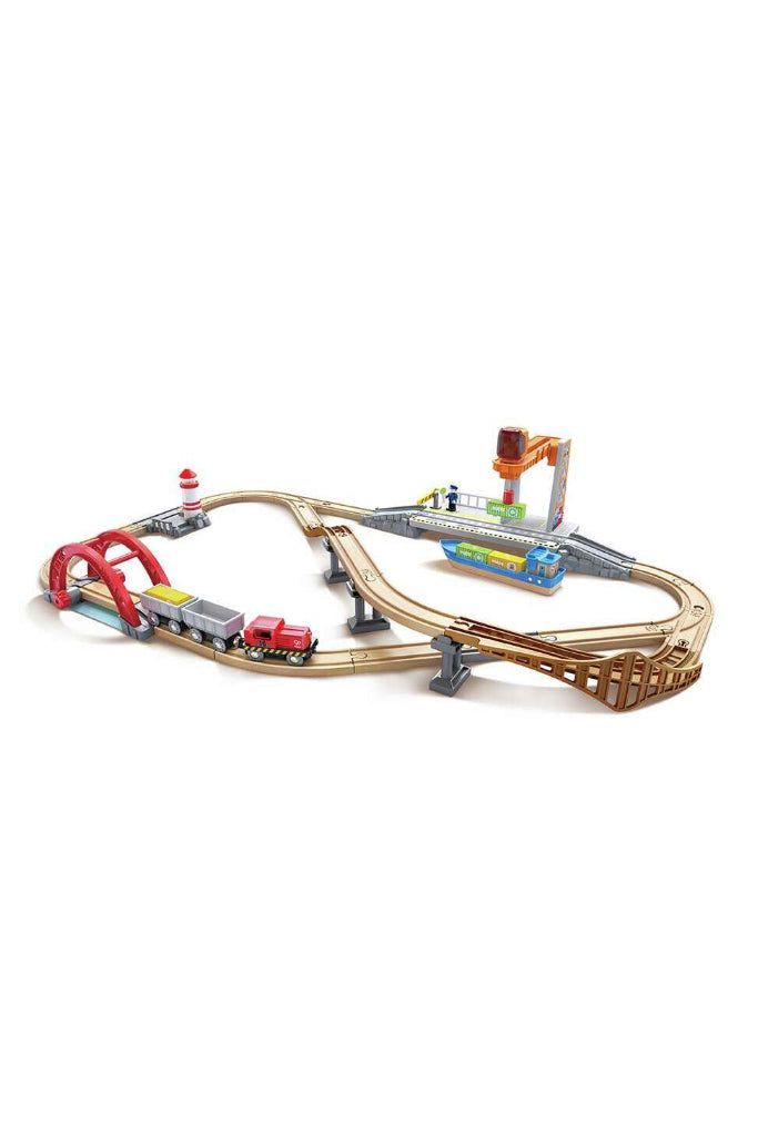 A toy train track set with curved and elevated wooden-style tracks, a red arched bridge, a lighthouse, a crane, and a small train on a plain white background.