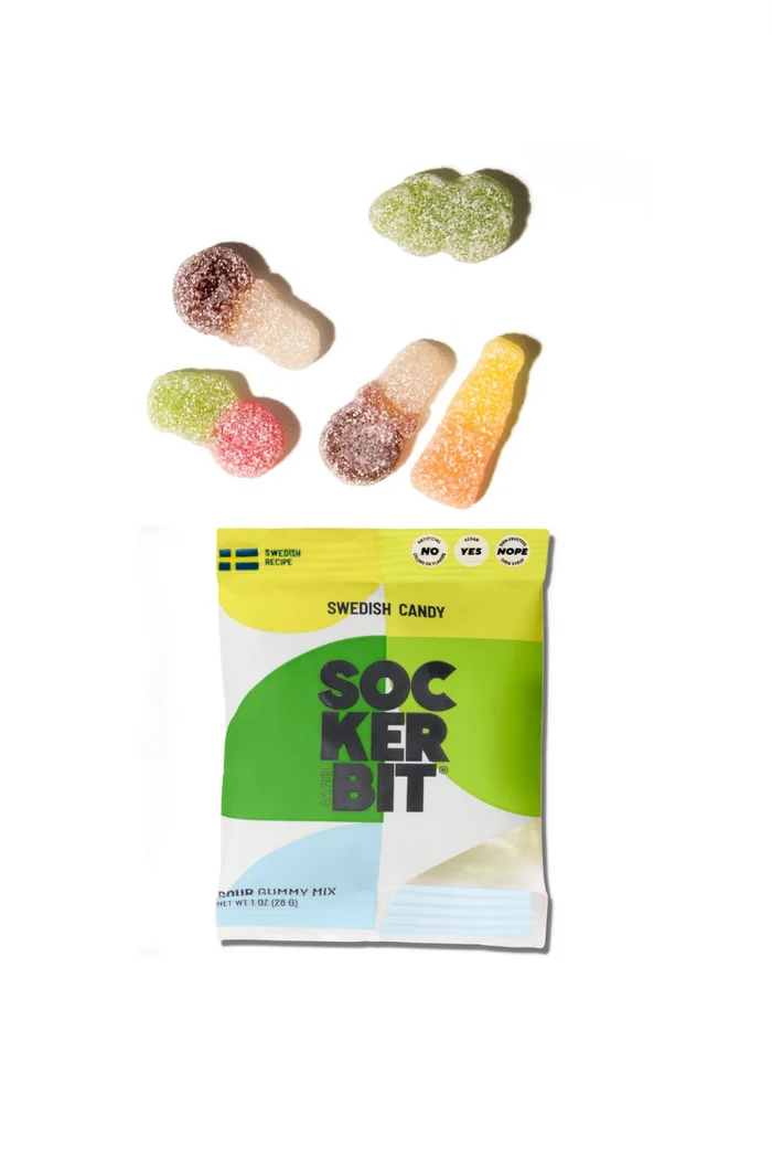 A bag of Swedish candy Sockerbit with several colorful, sugar-coated gummy candies scattered above it on a plain white background.