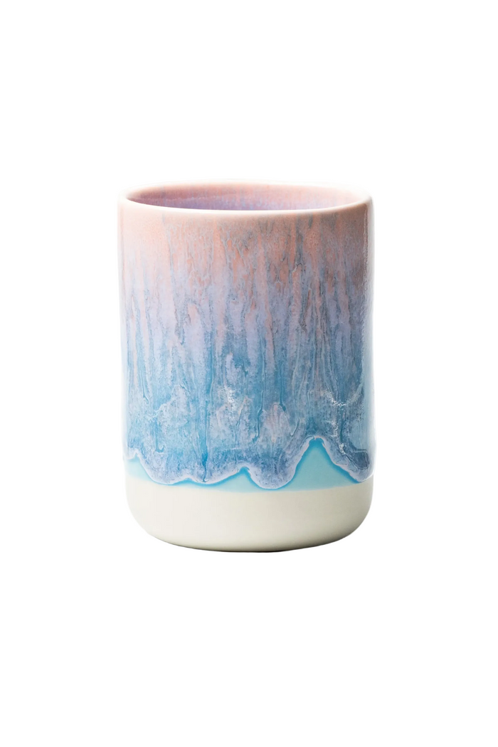 A ceramic cup with a dripping glaze design, featuring a pink and blue marbled pattern on the upper body and a solid cream-colored base, set against a plain white background.