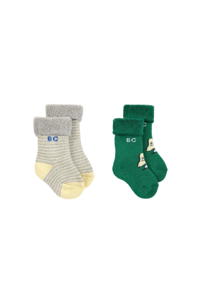 Two pairs of children's socks on a light gray background. The left pair is gray and white striped with a yellow toe and heel, featuring a fuzzy gray cuff and 'BC' in blue.