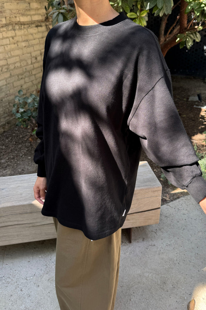 A person wearing a black oversized long-sleeve crewneck sweatshirt and beige pants, standing outdoors in front of a wooden bench with trees and a brick wall in the background.