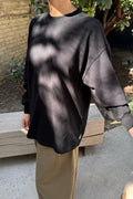 A person wearing a black oversized long-sleeve crewneck sweatshirt and beige pants, standing outdoors in front of a wooden bench with trees and a brick wall in the background.